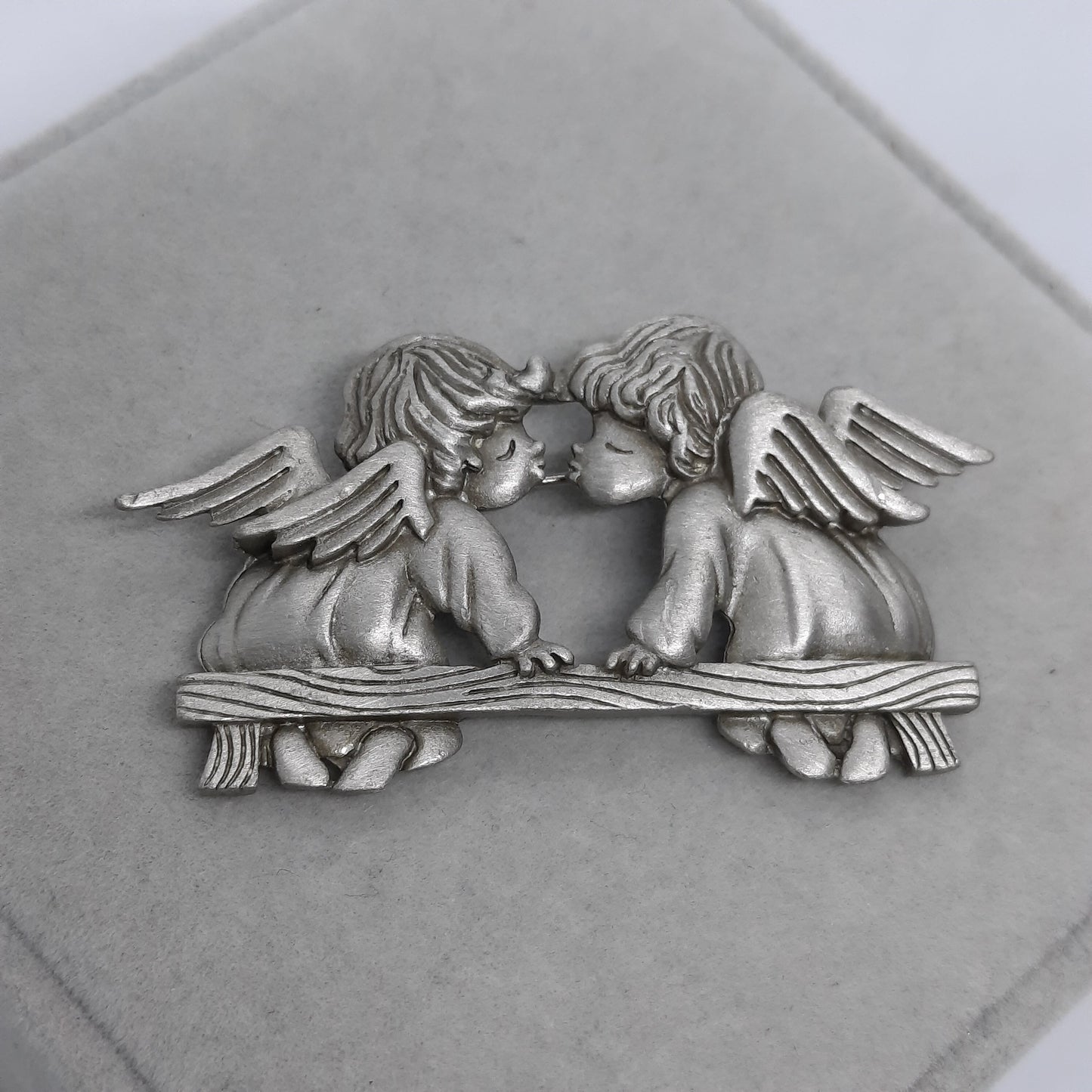 A silver metal brooch of two angels kissing. The angels are sitting on a bench with wings spread out behind them.