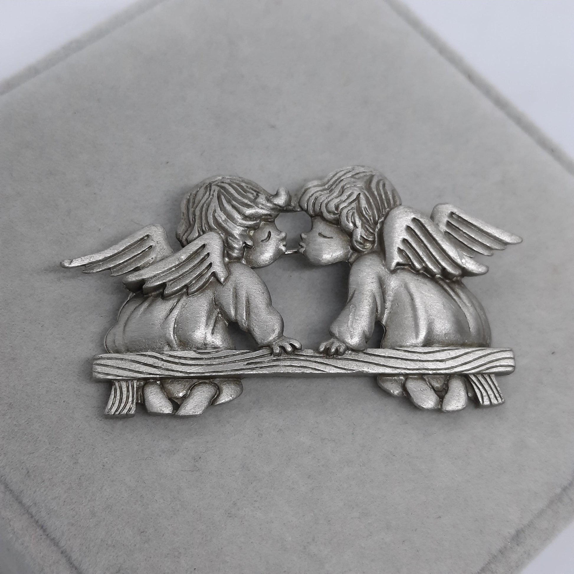 A silver metal brooch of two angels kissing. The angels are sitting on a bench with wings spread out behind them.