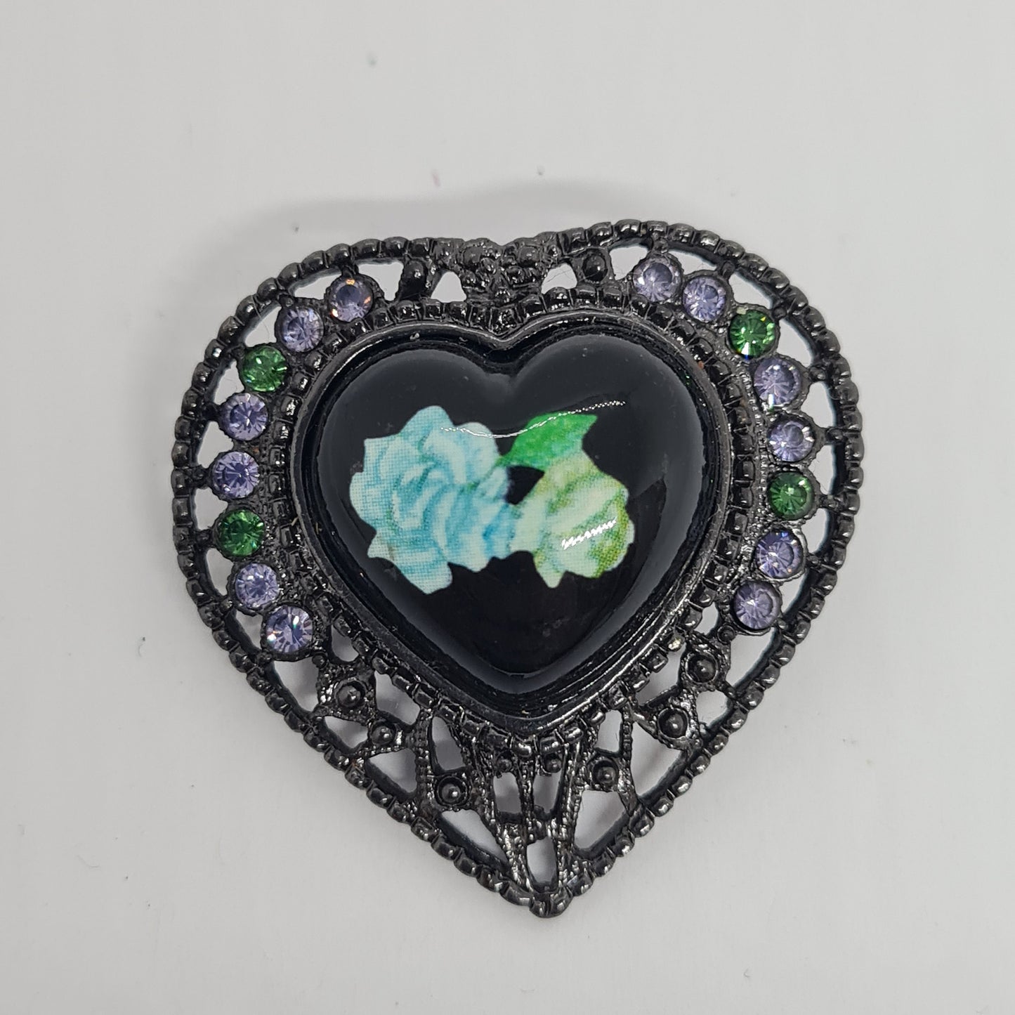 May include: A black heart-shaped pendant with a black enamel center featuring a blue and green floral design. The pendant is surrounded by a filigree design with purple and green rhinestones.