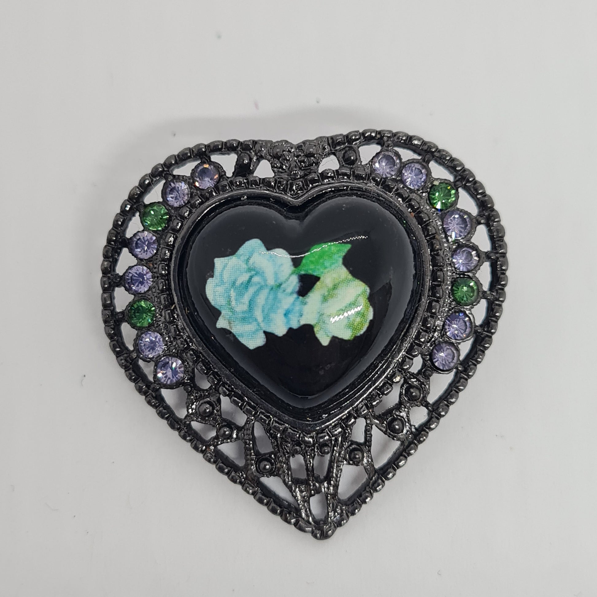 May include: A black heart-shaped pendant with a black enamel center featuring a blue and green floral design. The pendant is surrounded by a filigree design with purple and green rhinestones.